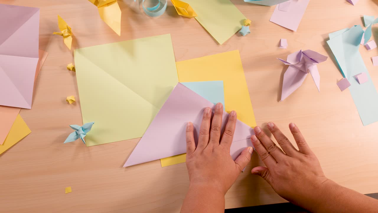 Person folds pastel origami paper on wooden table, surrounded by completed paper crafts, overhead view