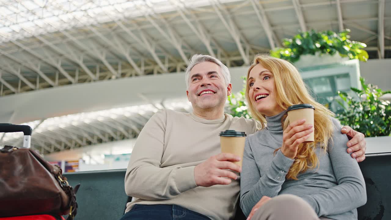 성숙 한 부부 가 공항 의 대기실 에 앉아서 커피 를 마시며 승객 들 에 대해 논의 하고 있다.