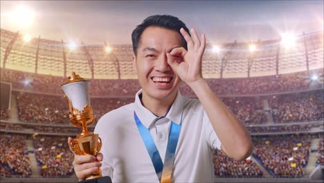 close up of asian man with a gold medal and trophy showing okay hand sign over eye and smiling to camera as the first winner at the stadium