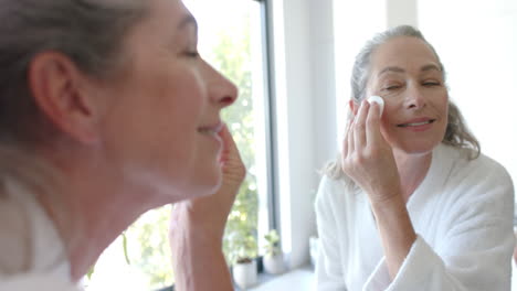 applying skincare, mature woman in bathrobe looking in mirror, smiling in bathroom