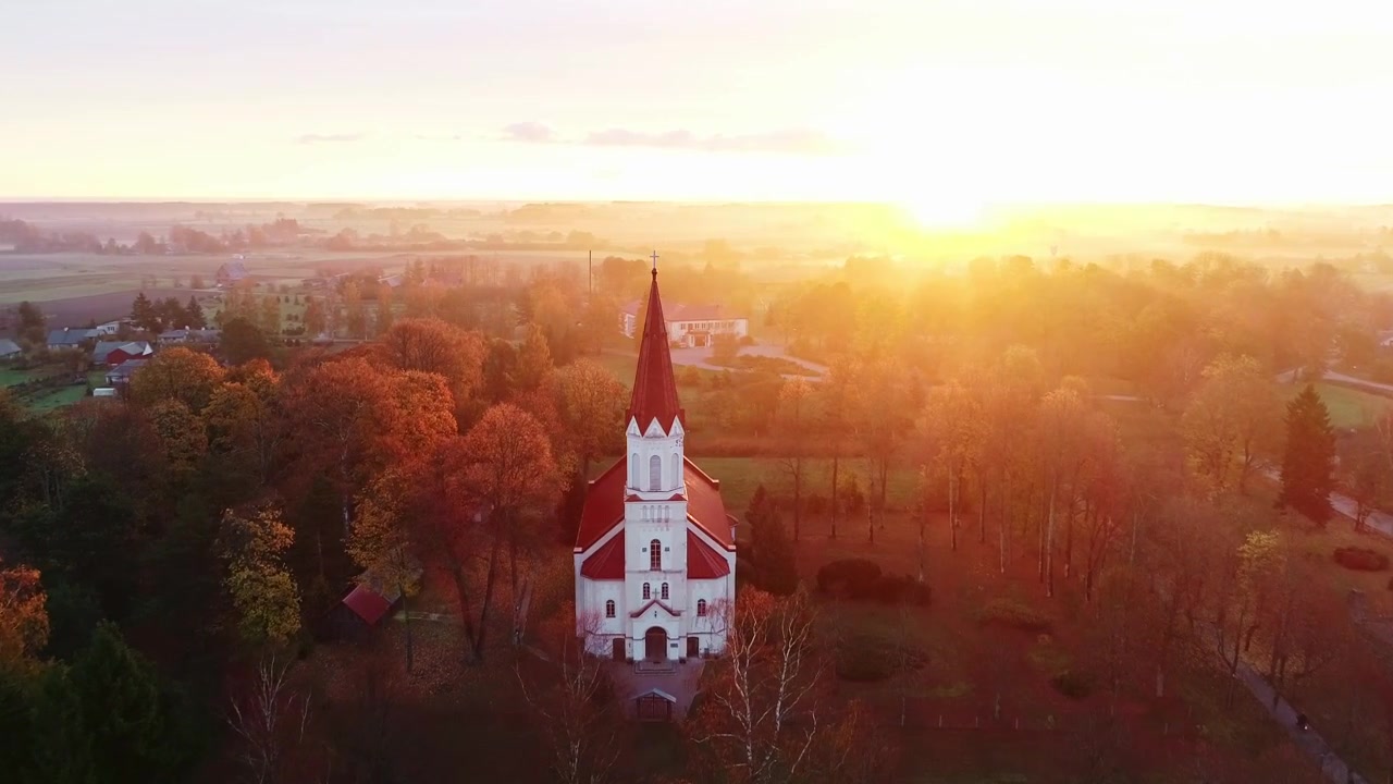 Lämmin auringonnousu kehystää Rucavan kirkon ja syksyisen maaseudun rauhallisessa ilmakuvassa.