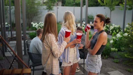 Three-happy-girl-friends-hold-red-glasses-in-their-hands-communicate-and-have-fun-while-relaxing-in-the-courtyard-of-a-country-house