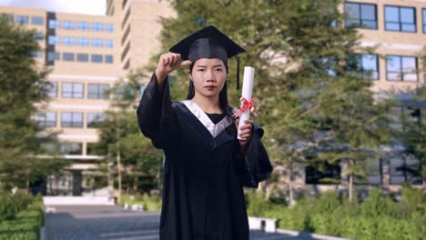 asian woman student graduates in cap and gown with diploma pointing to camera and showing thumbs down gesture in front of a magnificent university building