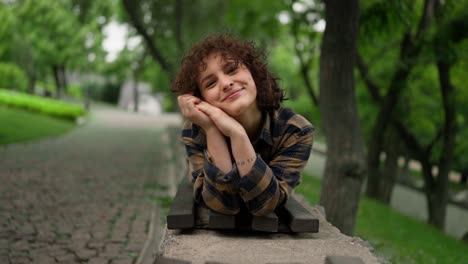 retrato de una estudiante feliz con el cabello rizado acostada en un banco y posando en el parque