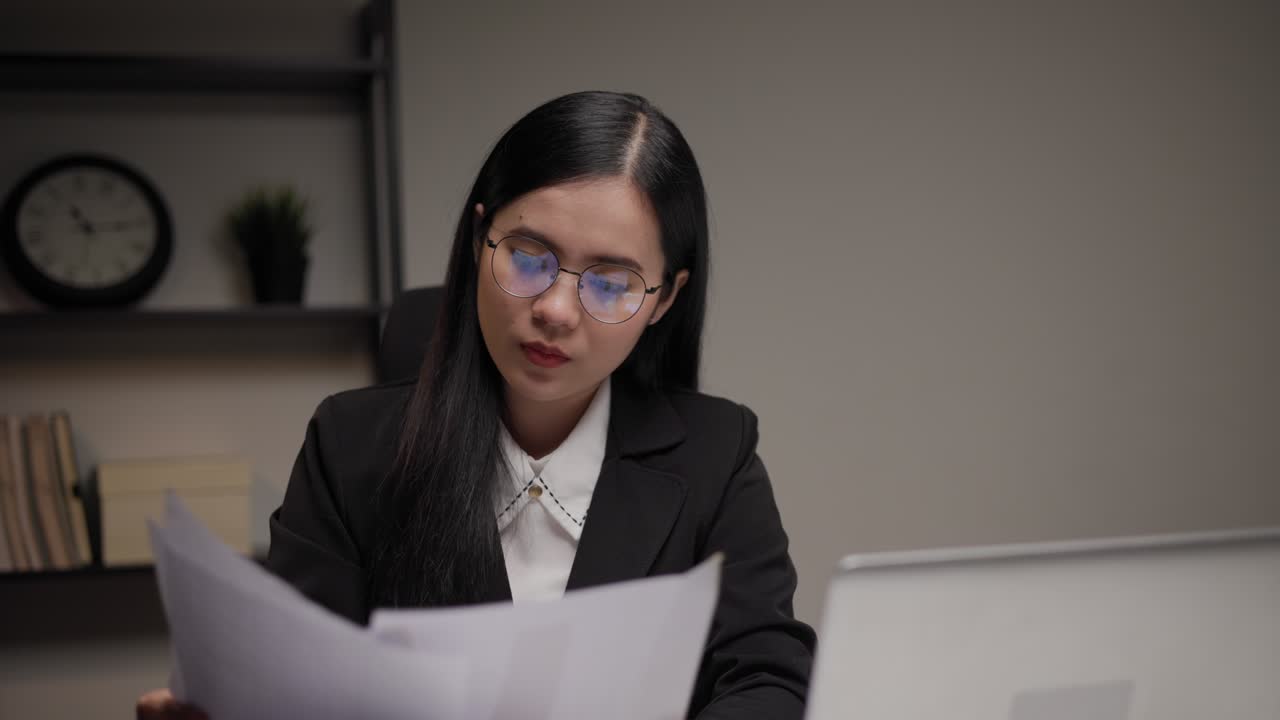 joven mujer de negocios asiática trabajando tarde en la noche. ella estaba muy ocupada comprobando el papeleo y tuvo que enviar el trabajo antes de la fecha límite, sintiéndose estresada. sentada en la oficina oscura por la noche.