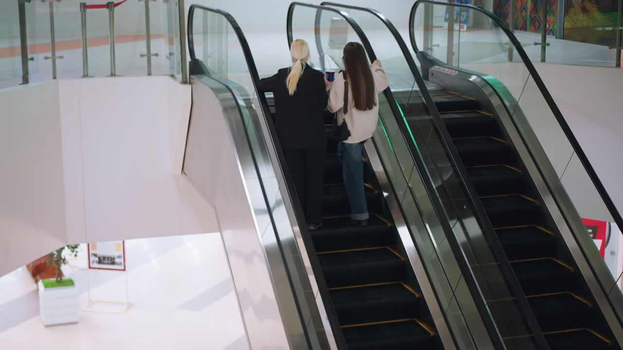 Fjern udsigt af to kvinder, der tager en rulletrappe op i et moderne indkøbscenter. Den ene holder en kop med en drink, mens de snakker afslappet og viser en afslappet livsstil og et åbenhjertigt, urbant samspil