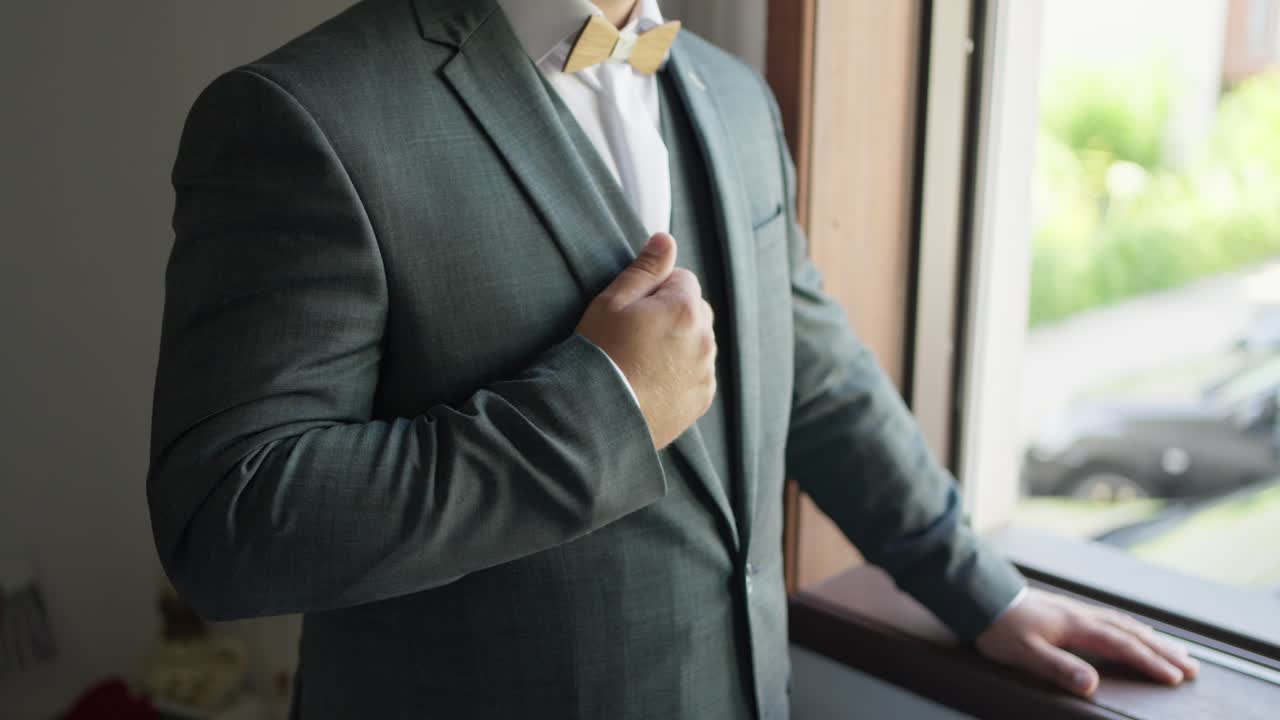 Primer plano del novio arreglándose el traje y la pajarita cerca de la ventana antes de la boda