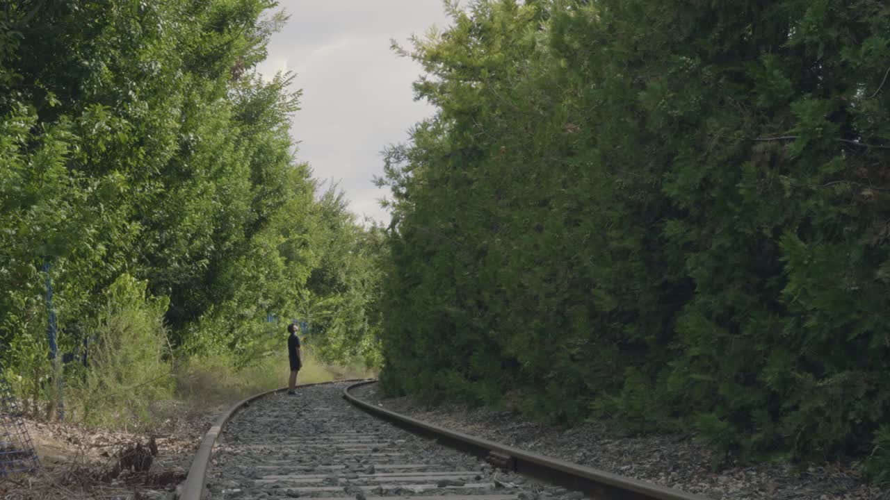 A man stands on abandoned railway tracks in the middle of a beautiful forest, admiring the surrounding nature. Ideal for travel, journey, reflection, freedom, or adventure in natural landscapes