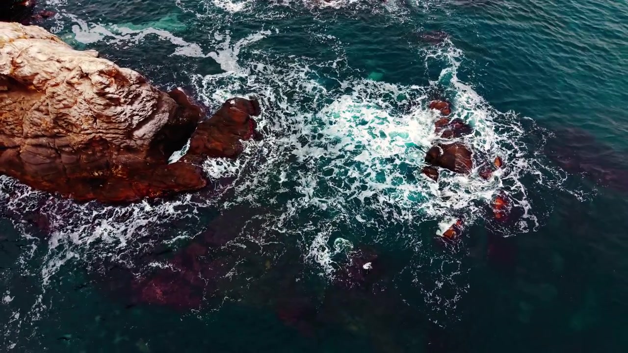 Ripresa aerea con drone, direttamente dall'alto, di spuma bianca che si infrange intorno a formazioni rocciose in acque color verde petrolio intenso. Onde marine che si infrangono su scogli costieri scuri, costa del Mar Nero.