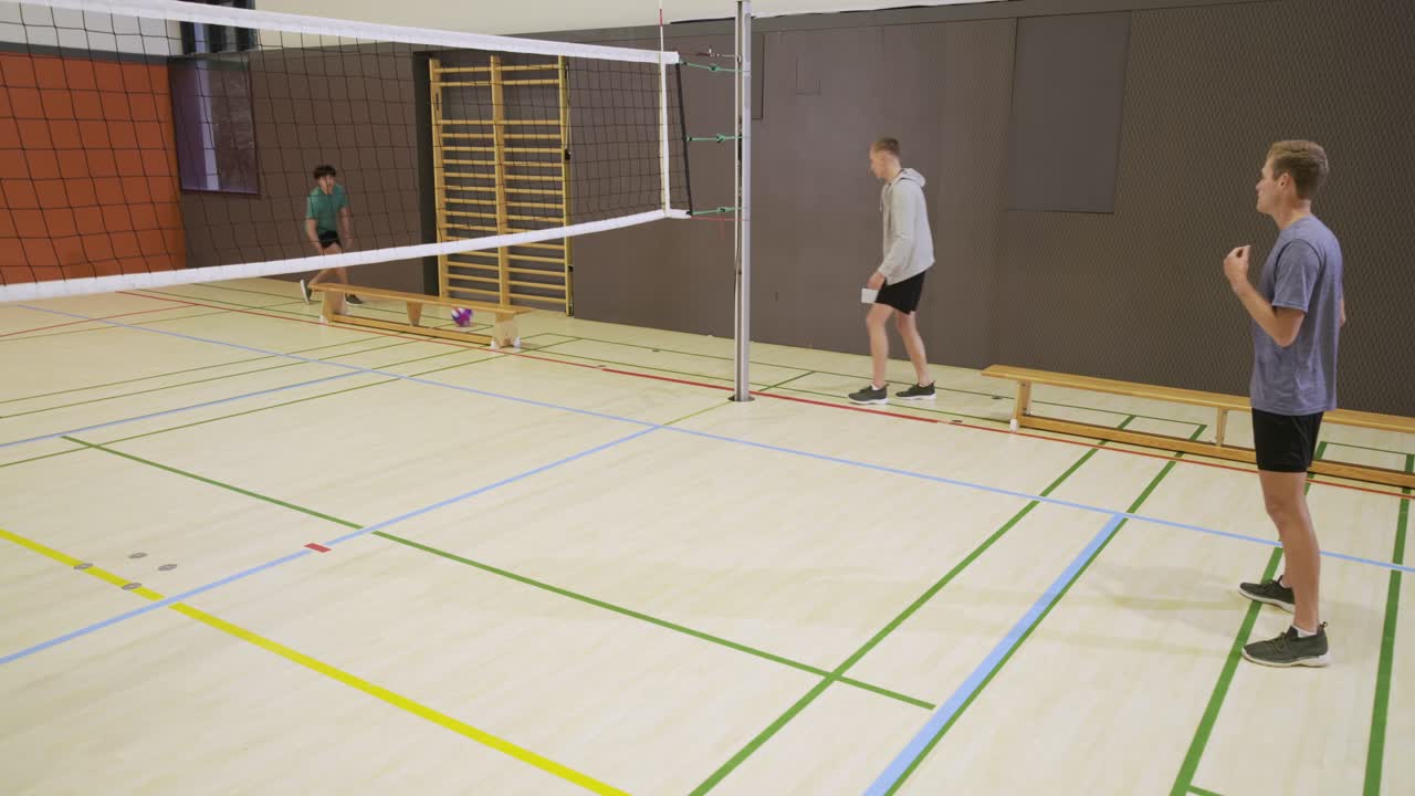Rozmanití muži hrají; muž v mikině přehazuje fialový volejbal přes síť a procvičují přihrávky v tělocvičně.