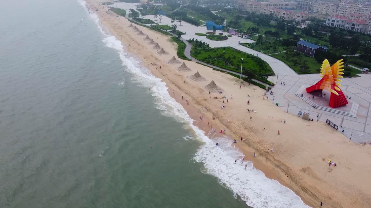 Aerial view flying from the sea toward sandy beach, bright red and yellow monument and sand sculptures