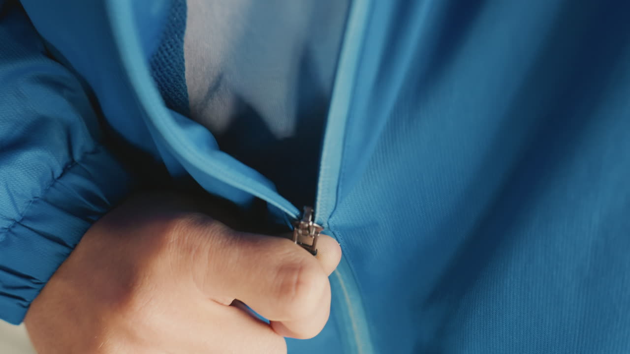 Closeup resleting jaket biru, tangan pelatih sedang bersiap. Momen pra-pertandingan hujan dengan pakaian luar nilon basah, patch reflektif, cengkeraman mantap pada slider, gerakan berirama saat pelatih mengamankan seragam sebelum pertandingan. Close
