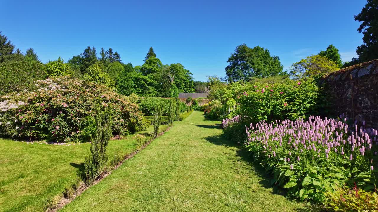 Smooth forward drone movement over a long garden pathway framed by lush hedges and flowering plants in Upper Brittany Botanical Park, Le Chatellier, France.