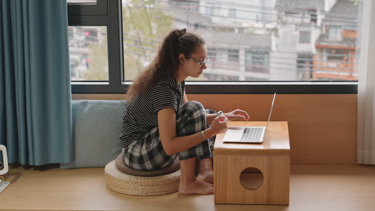 Mujer escribiendo en una laptop junto a la ventana, un rincón acogedor de hotel con vista al horizonte urbano, sentada en un cojín con pantalones de descanso a cuadros, postura relajada, luz natural y un ambiente tranquilo para el trabajo remoto.