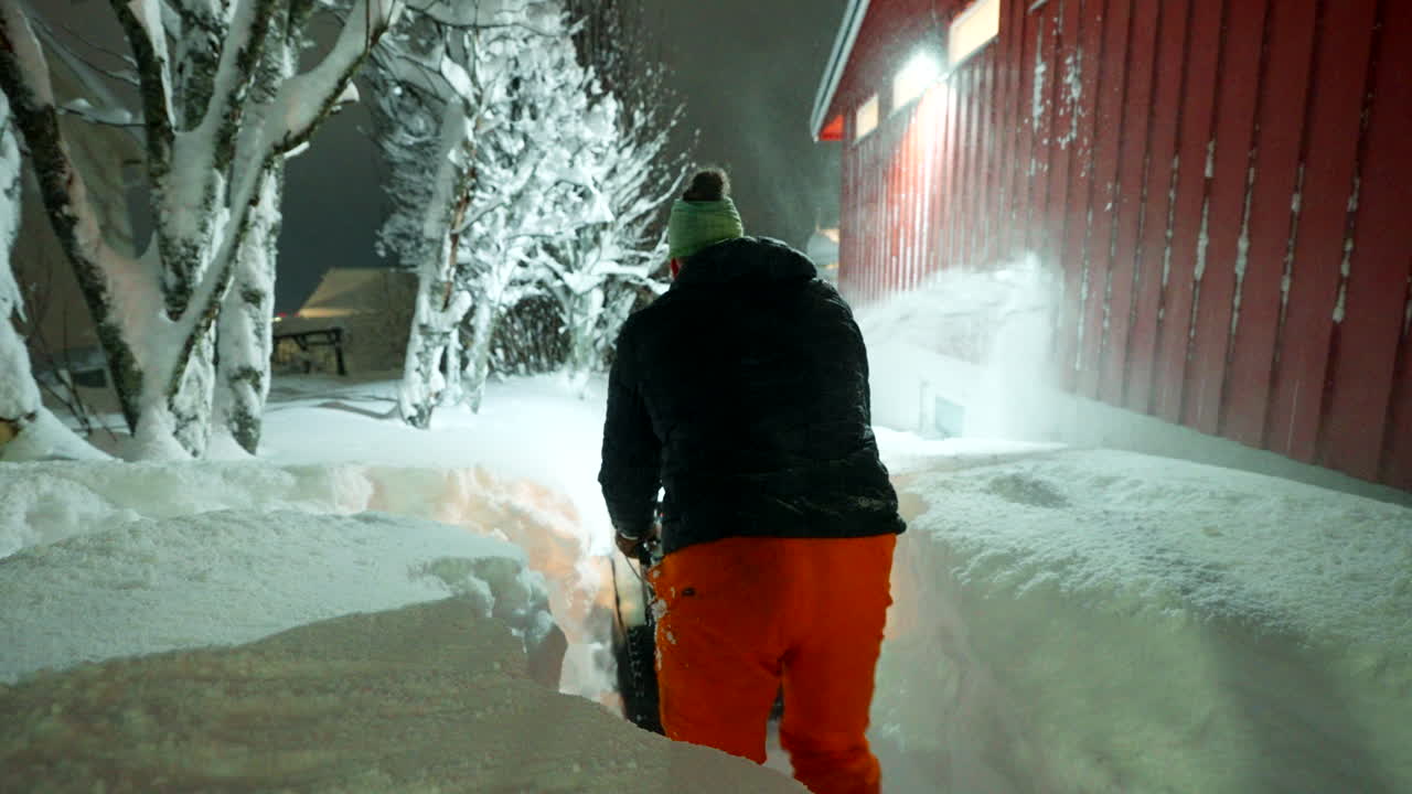 A lone man walks around corner clearing path in snowy Tromsø area around building as snowplow operate, arctic winter scene static