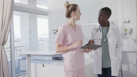 happy diverse female doctor and nurse in discussion in hospital ward using tablet, slow motion