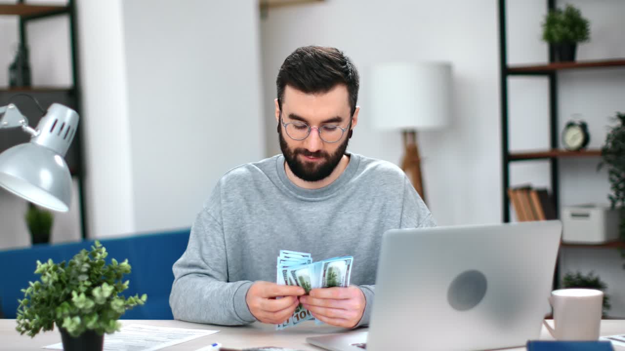 Hombre de negocios hipster exitoso y sonriente contando un fajo de dólares en su moderno lugar de trabajo con laptop.