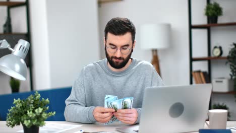 sonriente exitoso hombre de negocios hipster contando dinero en efectivo pila de dólares en el lugar de trabajo moderno portátil