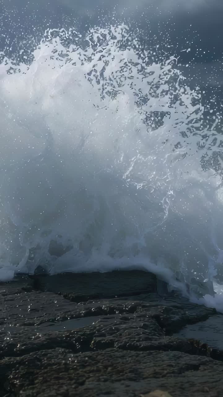 Vertical video: Surging ocean wave crashing onto dark rock ledge at shore, swell pushing foam