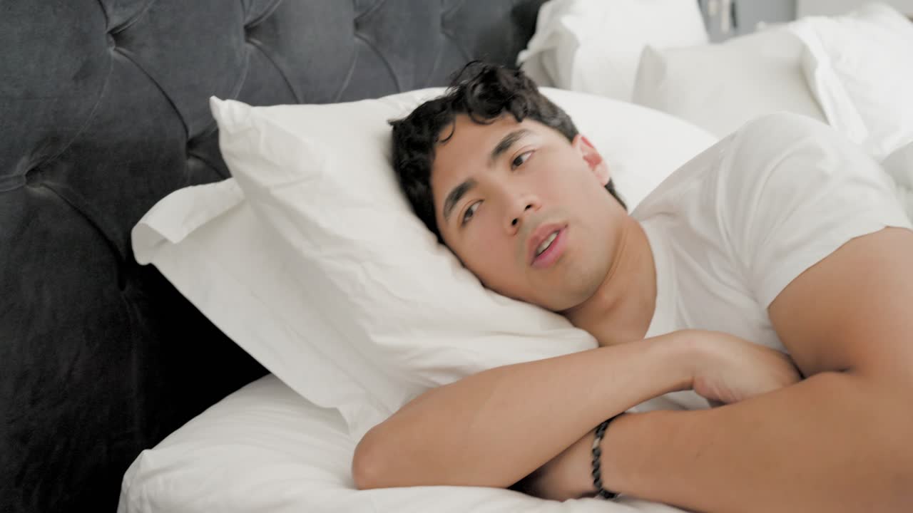 ベッド脇の電話で目覚め、手を伸ばしてスマートフォンをスクロールし、再び眠りに落ちるアジア人男性。