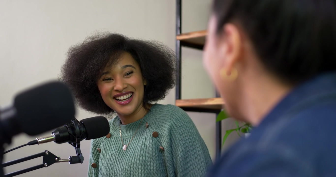 Forskellige kvindelige værter podcaster; den afroamerikanske vært smiler og læner sig mod mikrofonen, mens den asiatiske medvært taler.
