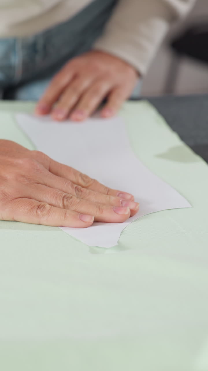 Mujer blanca colocando un patrón de papel sobre tela verde pálido, con alfileres y un marcador cerca, alineando y trazando cuidadosamente el patrón para cortar, con las manos alisando y preparando las piezas de la prenda en un taller ajetreado