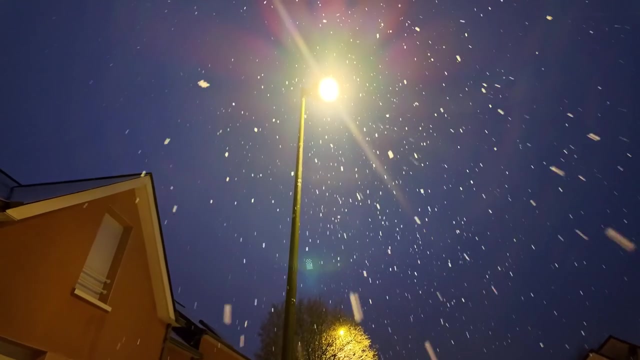 Poste de luz iluminando neve caindo à noite em bairro residencial.