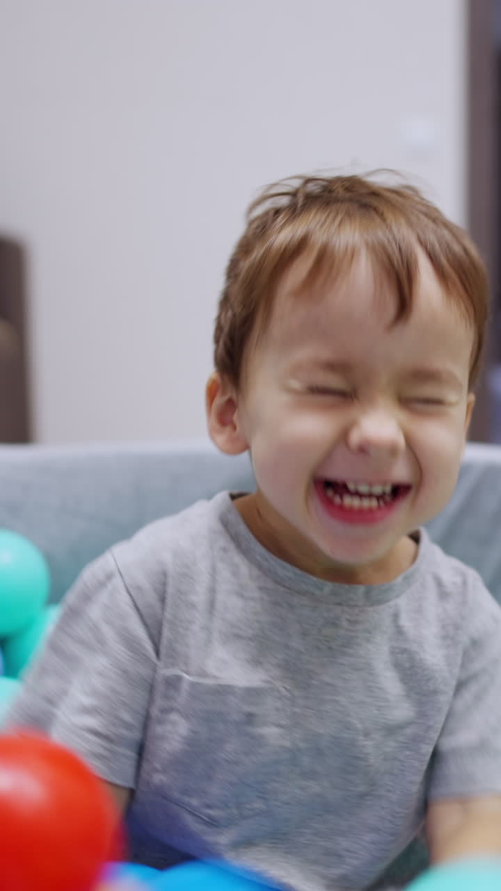 Lovely boy in grey t-shirt sits in the dry pool. Kid holds one blue ball on the head, drops it and waves hands to the sides. Vertical video