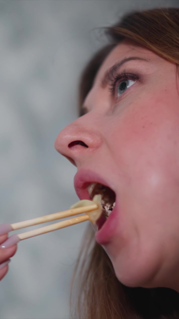 Close up of woman eating with chopsticks holding them near her mouth. scene is set against plain background with soft lighting focusing on action and texture details of food consumption