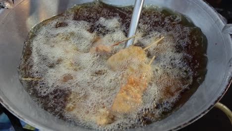 SLOW-MO:-High-angle-view-of-food-vendor-cooking-and-stirring-deep-fried-pork-on-stick-in-hot-boiling-oil-in-a-frying-pan-at-walking-street