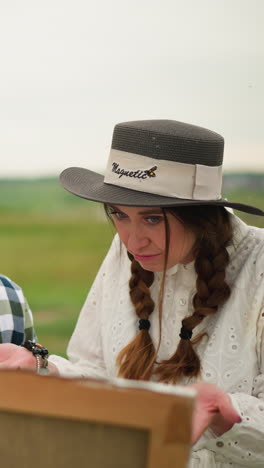an illustrator in a checkered shirt smiles as he works on his painting, set against a lush green landscape. beside him, a woman in a white dress and hat stands, observing his work with interest