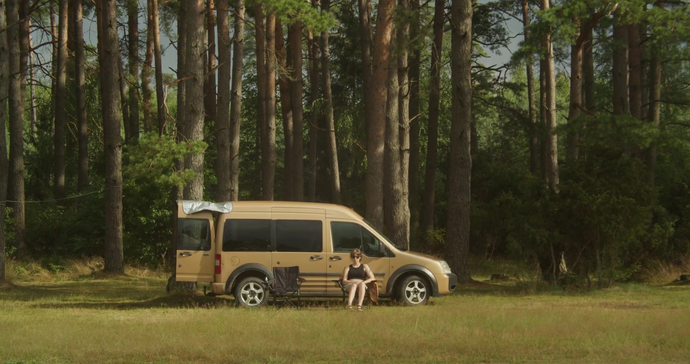 Una scena autentica di una nomade digitale seduta su una sedia fuori dal suo camper in una pineta illuminata dal sole. Perfetta per temi di van life, sostenibilità e lavoro da remoto.