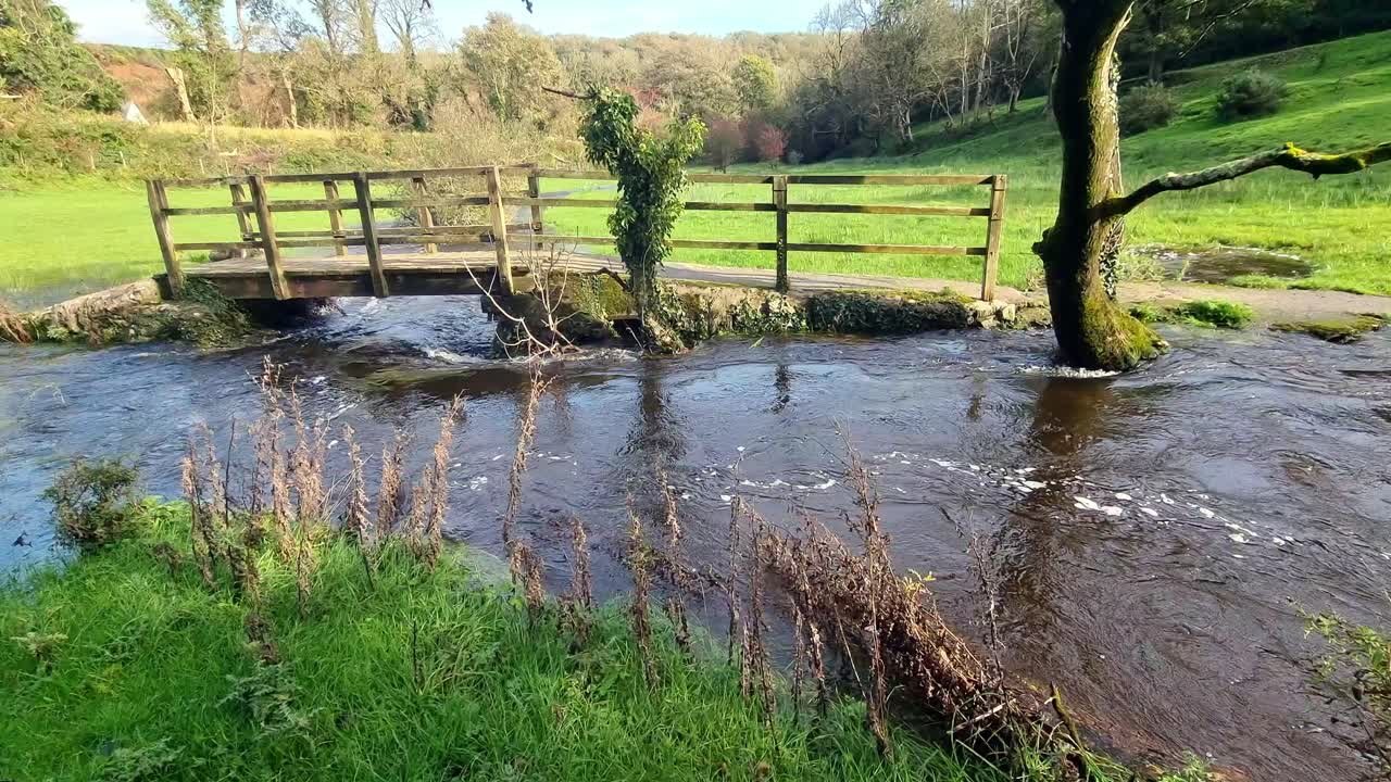 Overflowing burst riverbank flooding peaceful sunlit North Wales meadow under wooden bridge crossing