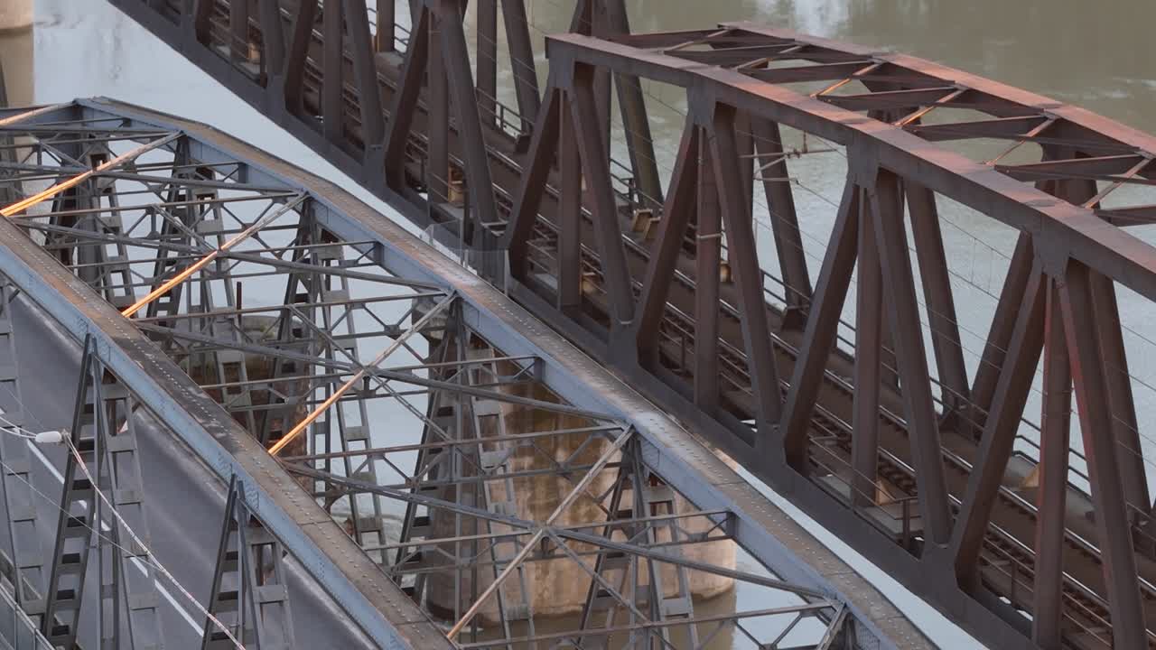 Drone ascends while tilting camera downward to reveal the detailed latticework of the historic iron bridge over the Po River near Cremona, surrounded by green trees and calm waters below