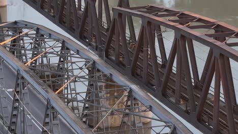 drone ascends while tilting camera downward to reveal the detailed latticework of the historic iron bridge over the po river near cremona, surrounded by green trees and calm waters below