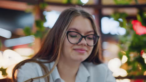 close-up of elegant lady wearing glasses, smiling while focusing on operating phone, warm bokeh lights in background enhance modern and stylish indoor atmosphere