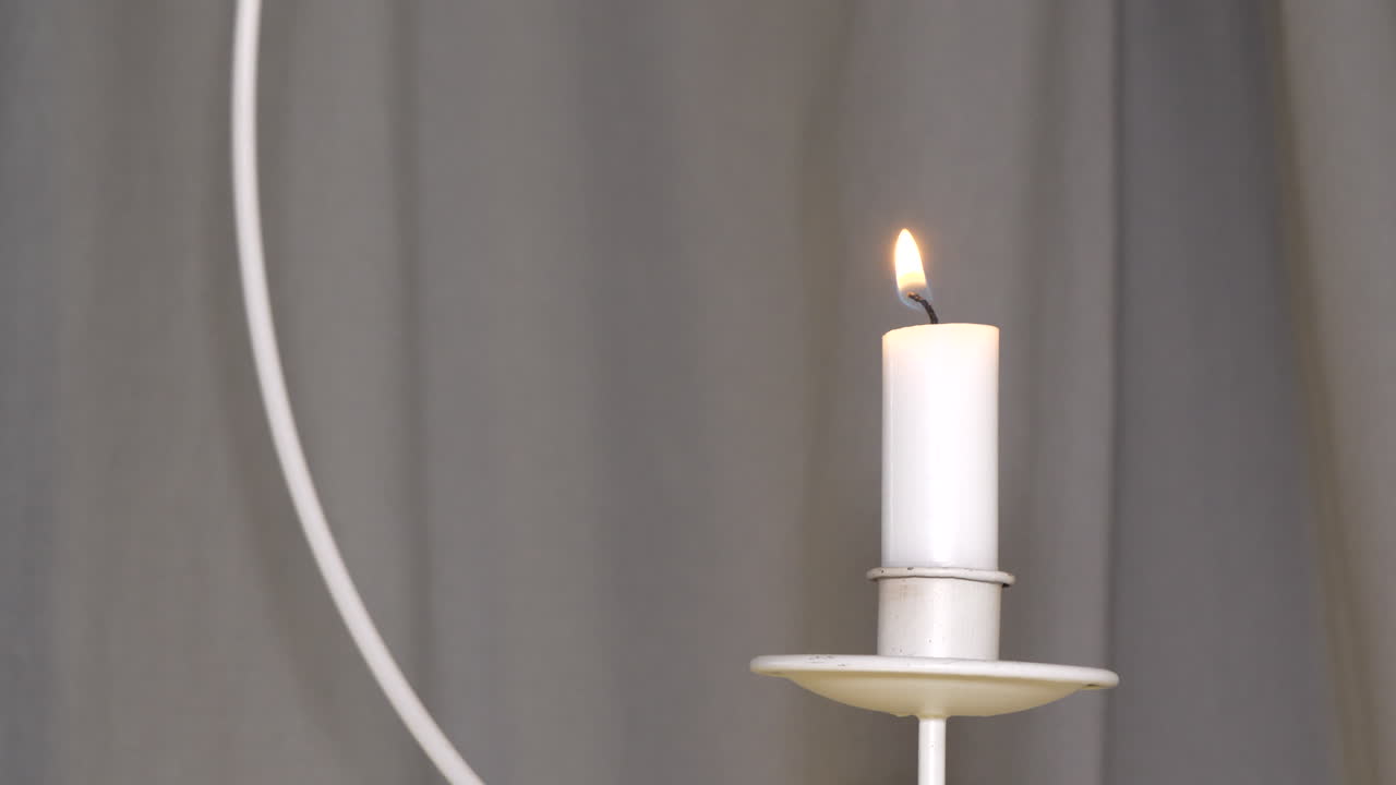 Nerozpoznatelná mužská ruka zapaluje visící svíčku na pozadí tmavého závěsu, kouř stoupá a rozptyluje se.