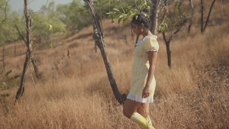 an attractive asian female wearing a pretty yellow checked dress dancing alone carefree around a tree in the middle of a remote dry grass field, india