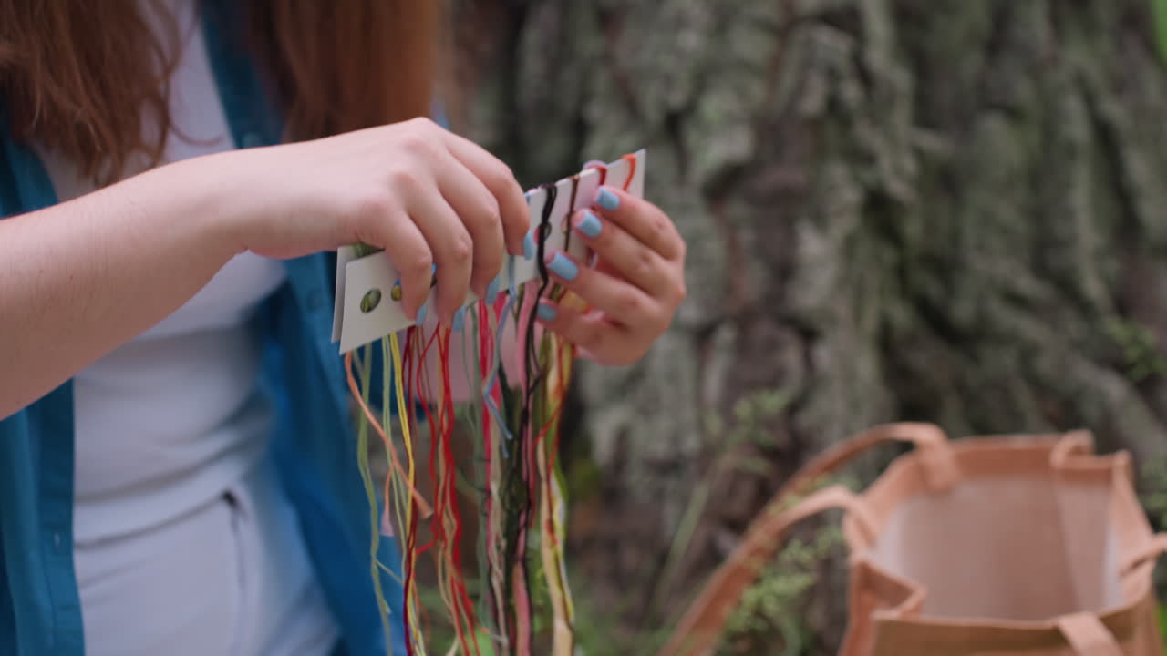 Primer plano de una mujer sosteniendo hilos de bordar de colores organizados en una tarjeta, sentada al aire libre cerca de un árbol, preparándose para una sesión de costura creativa en un entorno natural tranquilo y rodeado de luz solar.