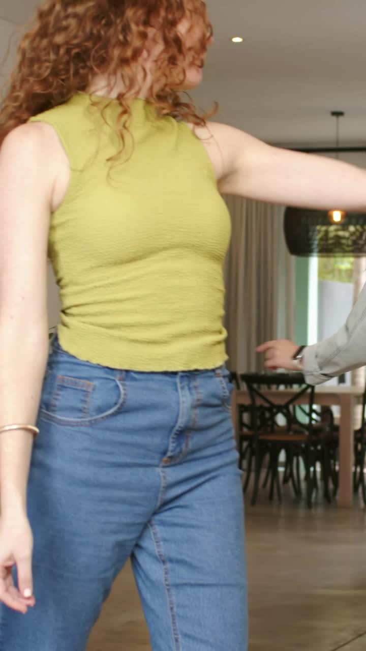 Video verticale: Con le mani alzate, una ragazza dai capelli rossi con un top verde che balla e trascina un amico verso una lampada in casa per divertimento.