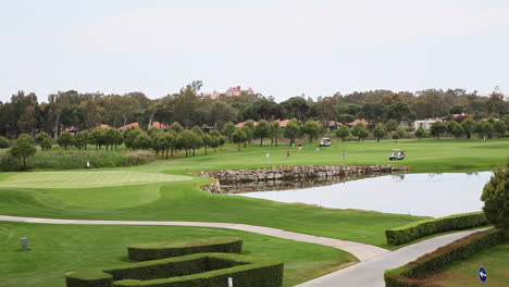 Timelapse-of-Playing-Golfers-on-Green-Fields-of-Golf-Course-with-Caddie-Carts,-Lakes-and-Trees-under-Open-Sky