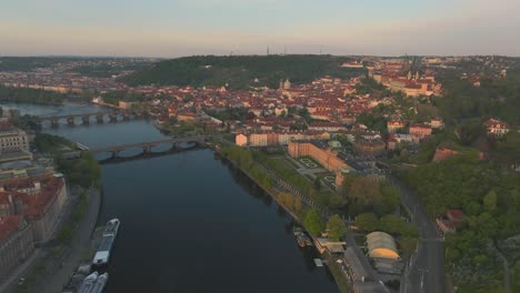 Drone-fly-along-Vlatva-River-in-Prague-City,-sailboats-docked,-green-hill-houses