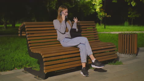 young lady seated on outdoor bench at night, multitasking with phone call and laptop, illuminated by soft park light, red backpack beside her on bench, surrounded by greenery