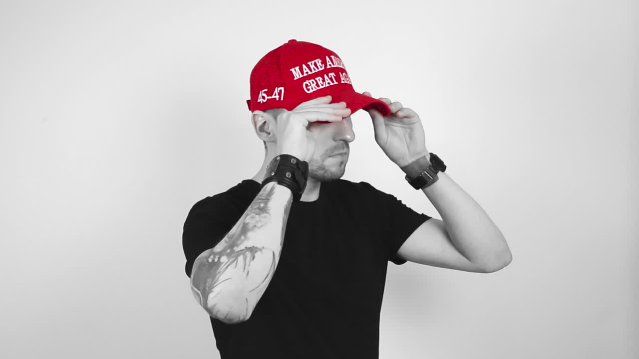 Grayscale footage of a tattooed and short bearded man putting on a vibrant colorful and red MAGA or Make America Great Again hat and looks directly into camera with a confident look and crossing arms.