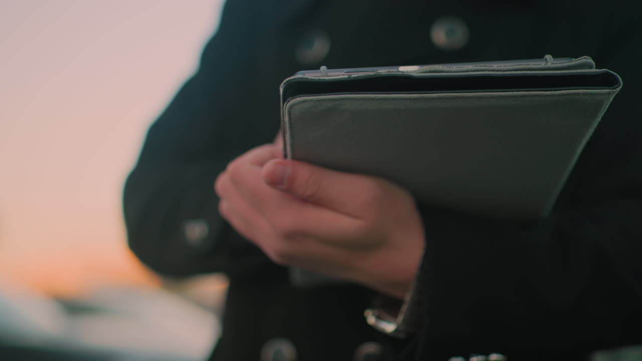 어두운 코트를 입은 개인의 클로즈업 태블릿을 운영하고, 어두운 밤에 배경에 흐릿한 인물이 있으며, 저녁 도시 분위기에서 기술에 참여하는 비즈니스 전문가를 포착합니다.