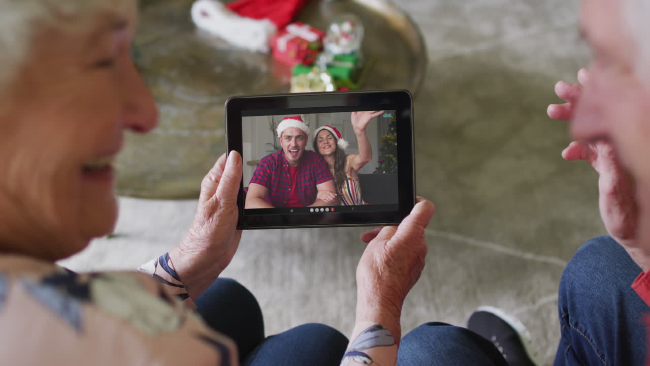 Anziana coppia caucasica che usa un tablet per una videochiamata natalizia con una coppia sorridente sullo schermo