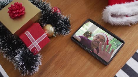 Smiling-biracial-man-wearing-santa-hat-on-christmas-video-call-on-tablet