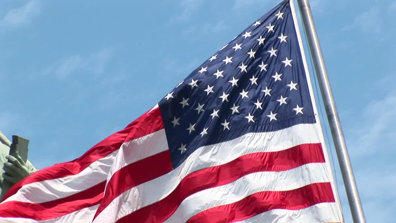 La telecamera esegue una panoramica a sinistra su una bandiera americana che ondeggia, rivelando la Statua della Libertà sullo sfondo.