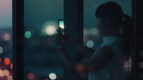 asian business woman using smartphone taking photo of city at night enjoying beautiful view working late in office