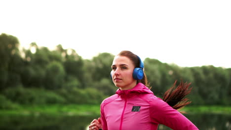 Ein-Mädchen-In-Rosa-Jacke-Und-Schwarzer-Hose-Läuft-Mit-Kopfhörern-Am-Fluss-Entlang-Und-Bereitet-Sich-Auf-Den-Marathon-Vor